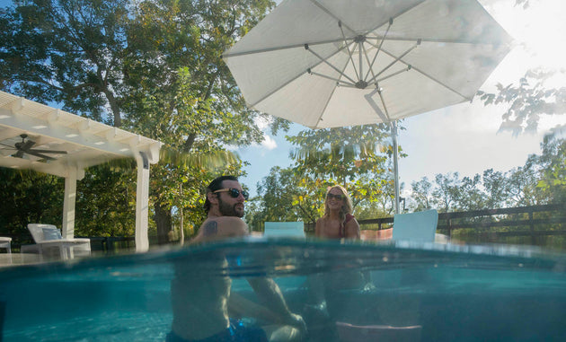 Two people sit in a hot tub with water at mid-level, revealing their upper bodies. A Ledge Lounger Ultra Cantilever Umbrella - 13 Octagon stands on the patio, surrounded by trees under a clear sky. Sunlight filters through foliage and umbrella light, creating a relaxed outdoor setting.