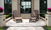 Two Legacy Adirondack by Ledge Lounger chairs with footrests sit on a patio in front of a modern building, ideal for gatherings. Behind them are large windows and stairs, while planters with purple flowers and trimmed hedges add to the inviting outdoor setup.