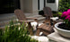 A pair of Legacy Adirondack Ottomans with white cushions by Ledge Lounger sit on a stone patio near a modern building. Nearby, theres a small table with a potted plant, and the pink flowers and green bushes create an ideal spot for outdoor relaxation.