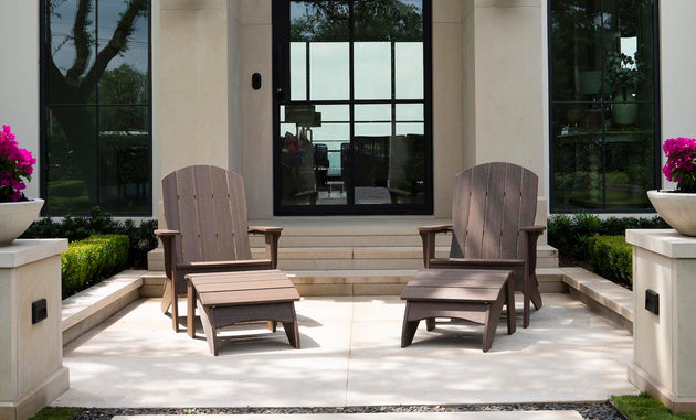 Outside a modern house with large glass doors, two Legacy Adirondack chairs and a matching Ledge Lounger Adirondack Ottoman create an inviting patio setup. The space, perfect for relaxation, features potted plants with purple blooms and trimmed greenery lining the steps.