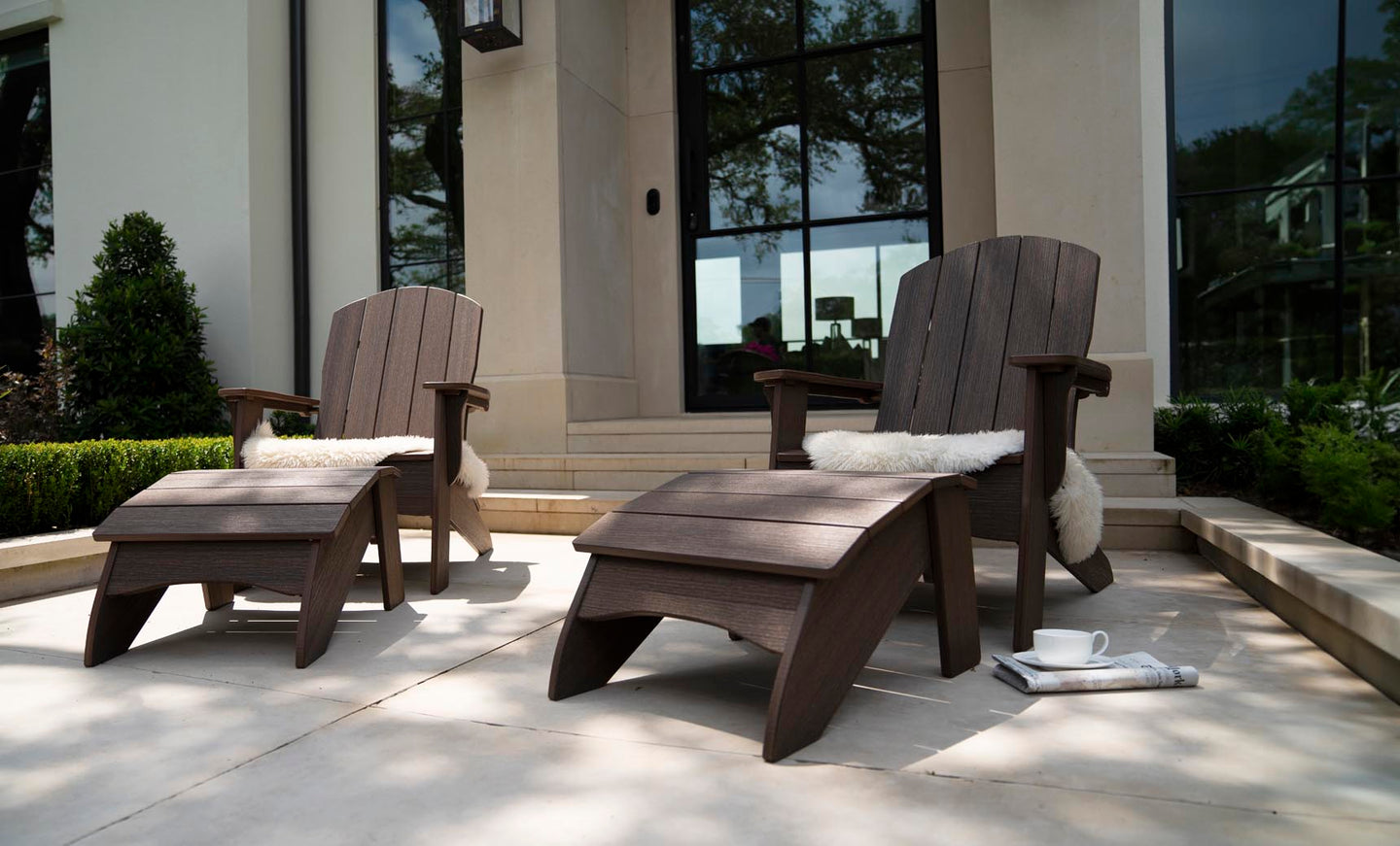 On a patio, two Ledge Lounger wooden Adirondack chairs with ottomans and sheepskin cushions offer outdoor relaxation. Nearby, a cup with saucer sits next to a folded newspaper. Surrounded by greenery, this tranquil setting is enhanced by the Legacy Adirondack Ottoman and a backdrop of glass doors.
