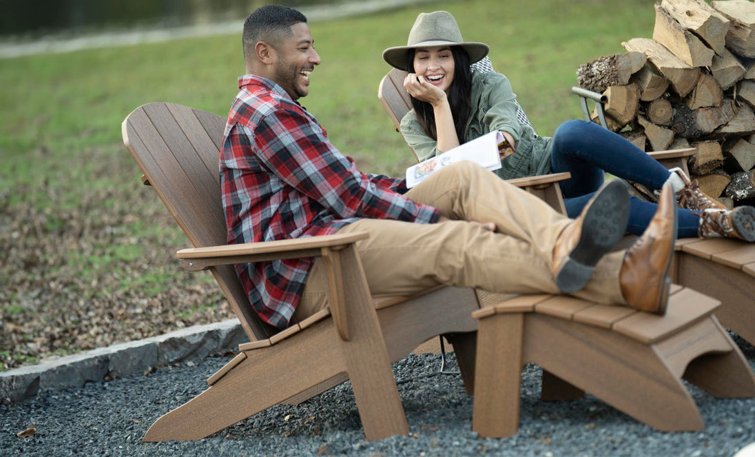A man in a red plaid shirt and a woman in a green jacket and hat sit outdoors in Legacy Adirondack chairs by a woodpile. Shes holding a magazine, both smiling, embodying outdoor relaxation with Ledge Loungers Legacy Adirondack Ottoman.