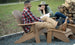 A man in a red plaid shirt and a woman in a green jacket and hat sit outdoors in Legacy Adirondack chairs by a woodpile. Shes holding a magazine, both smiling, embodying outdoor relaxation with Ledge Loungers Legacy Adirondack Ottoman.
