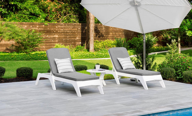 Two gray lounge chairs with striped outdoor fabric pillows sit beside a small round table under a large white umbrella on a tiled poolside patio, surrounded by a wooden fence, greenery, and trees—ideal for relaxation with Ledge Loungers customizable Legacy Chaise Cushion.
