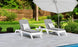 Two gray lounge chairs with striped outdoor fabric pillows sit beside a small round table under a large white umbrella on a tiled poolside patio, surrounded by a wooden fence, greenery, and trees—ideal for relaxation with Ledge Loungers customizable Legacy Chaise Cushion.