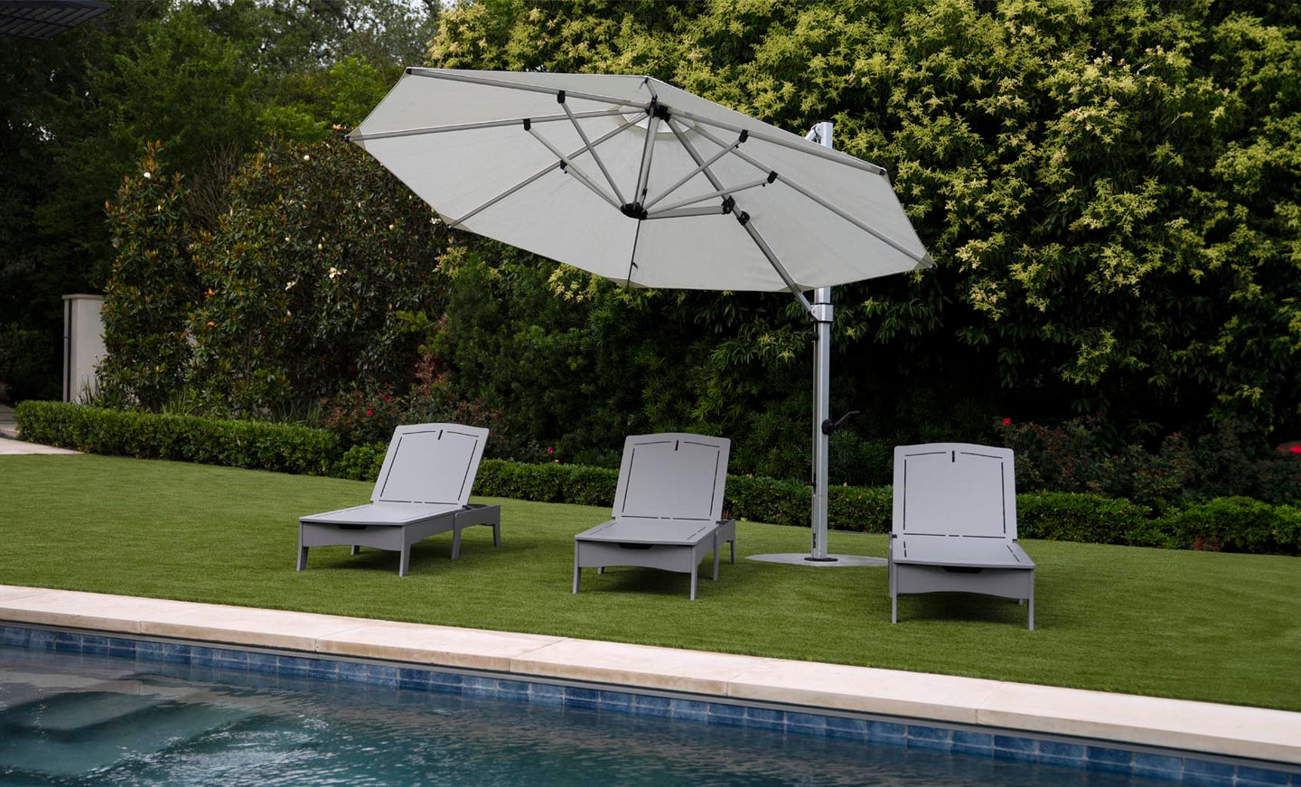 Three Ledge Lounger Legacy Chaise lounge chairs are set on green grass under a large white umbrella, adjustable to various reclining positions and facing the pool. Dense greenery with tall bushes and trees provides a serene backdrop to this tranquil outdoor scene.