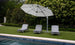 Three Ledge Lounger Legacy Chaise lounge chairs are set on green grass under a large white umbrella, adjustable to various reclining positions and facing the pool. Dense greenery with tall bushes and trees provides a serene backdrop to this tranquil outdoor scene.