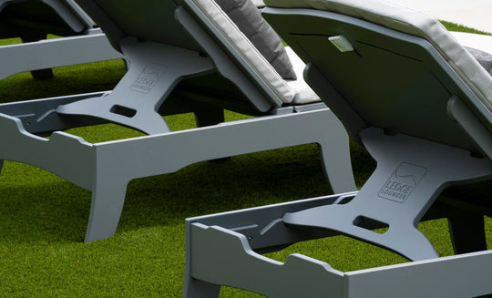 Close-up of grey Legacy Chaise lounge chairs from Ledge Lounger, lined up on green artificial turf. Featuring white cushions and multiple reclining positions, LEDGE LOUNGER is visible on the bases in this inviting outdoor space.