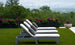 Three white Legacy Chaise Cushions by Ledge Lounger with gray pillows rest on grass, overlooking a misty cityscape. Behind them, planters with vibrant purple flowers and lush greenery add charm. The durable outdoor fabric offers customization to match any style.