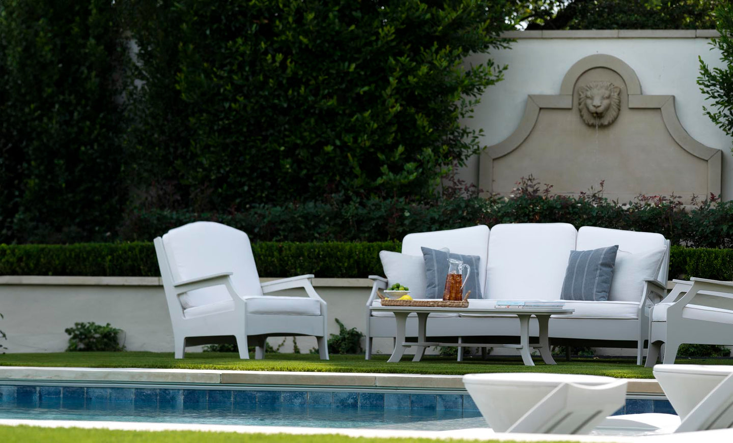 A serene patio scene features the elegant Legacy Sofa by Ledge Lounger, with gray and white outdoor fabrics by the pool. An armchair complements the setting, surrounded by lush greenery. A decorative wall with a lion relief adds a touch of elegance to this tranquil oasis.
