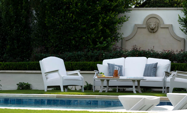 A serene patio scene features the elegant Legacy Sofa by Ledge Lounger, with gray and white outdoor fabrics by the pool. An armchair complements the setting, surrounded by lush greenery. A decorative wall with a lion relief adds a touch of elegance to this tranquil oasis.