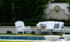 A serene patio scene features the elegant Legacy Sofa by Ledge Lounger, with gray and white outdoor fabrics by the pool. An armchair complements the setting, surrounded by lush greenery. A decorative wall with a lion relief adds a touch of elegance to this tranquil oasis.