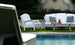 The serene poolside patio is furnished with elegant white lounge pieces, including a Legacy Club Chair by Ledge Lounger and a two-seater sofa. A table with iced tea adds to the scene, set against lush greenery and hedges for ultimate relaxation.