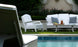The serene poolside patio is furnished with elegant white lounge pieces, including a Legacy Club Chair by Ledge Lounger and a two-seater sofa. A table with iced tea adds to the scene, set against lush greenery and hedges for ultimate relaxation.