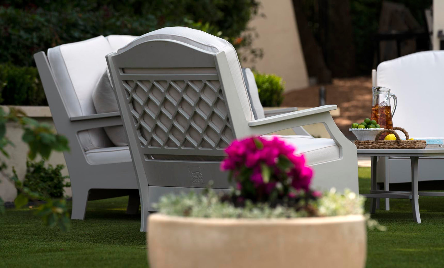 A cozy outdoor space boasts a pair of plush Legacy Club Chairs by Ledge Lounger, positioned around a low table with iced tea, glasses, grapes, and lemons. A large planter filled with vibrant pink flowers completes this inviting scene.