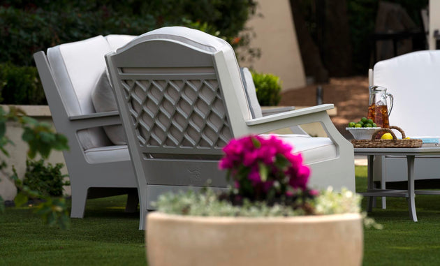 A cozy outdoor space boasts a pair of plush Legacy Club Chairs by Ledge Lounger, positioned around a low table with iced tea, glasses, grapes, and lemons. A large planter filled with vibrant pink flowers completes this inviting scene.