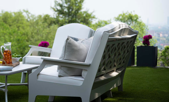 Legacy Club Chairs with cushions face a white coffee table on a grass lawn. A pitcher of iced tea and a snack bowl rest on the table. Potted purple flowers are nearby, framing the inviting outdoor space with blurred trees and a cityscape in the background. Brand: Ledge Lounger.