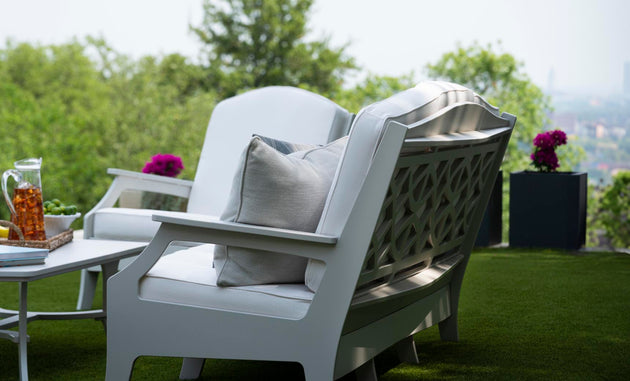 Legacy Club Chairs with cushions face a white coffee table on a grass lawn. A pitcher of iced tea and a snack bowl rest on the table. Potted purple flowers are nearby, framing the inviting outdoor space with blurred trees and a cityscape in the background. Brand: Ledge Lounger.