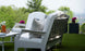 Legacy Club Chairs with cushions face a white coffee table on a grass lawn. A pitcher of iced tea and a snack bowl rest on the table. Potted purple flowers are nearby, framing the inviting outdoor space with blurred trees and a cityscape in the background. Brand: Ledge Lounger.