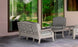 The outdoor patio features a Ledge Loungers Legacy Sofa and two chairs with durable white cushions. A coffee table sits on the tiled floor, while large glass windows reflect the surrounding greenery, enhancing the serene atmosphere.