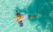 A blonde person in a blue swimsuit relaxes on the Luxurious Laze-y Days Float Bundle by Ledge Lounger in clear turquoise water, with sunlight dancing across the surface creating a serene tropical vibe.