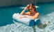 A woman in sunglasses and a pink bikini relaxes on a Laze Hammock by Ledge Lounger in a pool, with sunlight glistening on the water and a small fountain in the background.