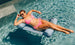 Wearing a pink bikini and sunglasses, a person relaxes on the Laze Hammock by Ledge Lounger in clear blue water. Theyre smiling, resting one hand on their sunglasses, and enjoying their sunny outdoor adventure.