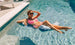 A person in sunglasses and a pink bikini relaxes on a Ledge Lounger Laze Hammock float in the sunlit pool, smiling with their arm resting on their head. The water is clear, and theres a white chair on the pool deck.