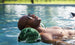 A man in sunglasses relaxes in a Ledge Lounger Laze Hammock float made with marine-grade fabrics, drifting in a calm pool. His eyes are closed and head tilted back as the water blends seamlessly with the blurred trees and grass around him.