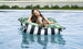 A woman relaxes contentedly on a Laze Pillow by Ledge Lounger in a pool, resting her head on her hand. The floating bean bag features black and white stripes with green leaf patterns along the edges, delivering ultimate outdoor comfort.