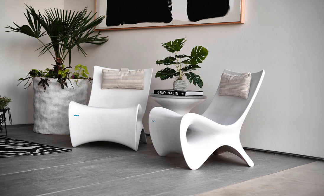 A modern interior features two sleek white chairs with beige cushions, centered around a small white table holding a stack of books and a potted plant. Large potted greenery sits nearby, and an abstract black-and-white artwork hangs above.