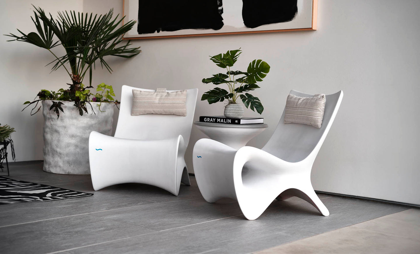 A modern interior features two sleek white chairs with beige cushions, centered around a small white table holding a stack of books and a potted plant. Large potted greenery sits nearby, and an abstract black-and-white artwork hangs above.