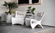 A modern interior features two sleek white chairs with beige cushions, centered around a small white table holding a stack of books and a potted plant. Large potted greenery sits nearby, and an abstract black-and-white artwork hangs above.