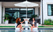 Two women in swimsuits enjoy outdoor moments on modern Autograph Chairs by Ledge Lounger. They clink glasses under a large white umbrella beside a sleek table, with a backdrop of luxury including a modern house with glass doors and elegant potted plants.