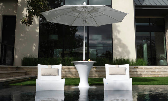Two Ledge Lounger Autograph Chairs with beige pillows face a sleek white table under a large umbrella outdoors, set by reflective water and enhanced by a contemporary building window in the serene backdrop.