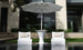 Two Ledge Lounger Autograph Chairs with beige pillows face a sleek white table under a large umbrella outdoors, set by reflective water and enhanced by a contemporary building window in the serene backdrop.
