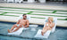 A man and woman enjoy a luxurious lounging experience on partially submerged Ledge Lounger Autograph Chaises. Clad in swimsuits and sunglasses, they smile over a small round table, with a modern patio enhancing the serene outdoor backdrop.