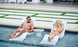 A man and woman enjoy a luxurious lounging experience on partially submerged Ledge Lounger Autograph Chaises. Clad in swimsuits and sunglasses, they smile over a small round table, with a modern patio enhancing the serene outdoor backdrop.
