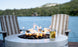 In the foreground, a round fire pit with flames is encircled by smooth stones. A glass mug containing a hot beverage rests on its edge. Two striped deck chairs with Ledge Lounger’s Mainstay Adirondack Cushion for comfort overlook a serene lake and lush green hills.