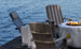 Three Adirondack chairs with striped cushions surround a fire pit on a dock by the lake. Each chair has a Mainstay Adirondack Headrest Pillow from Ledge Lounger, featuring high-quality fabric and an adjustable strap, with drinks in cup holders. A folded blanket rests on one chair while water ripples below.