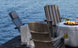 Three Adirondack chairs with striped cushions surround a fire pit on a dock by the lake. Each chair has a Mainstay Adirondack Headrest Pillow from Ledge Lounger, featuring high-quality fabric and an adjustable strap, with drinks in cup holders. A folded blanket rests on one chair while water ripples below.