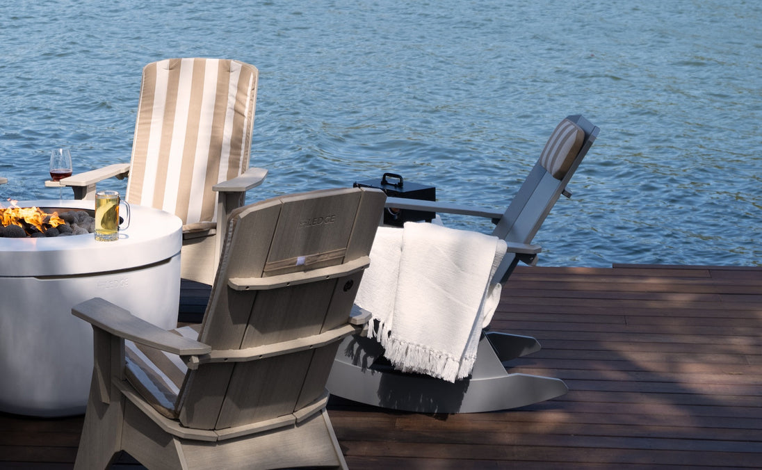 Two Mainstay Adirondack Rockers by Ledge Lounger on a wooden deck face serene water. A nearby fire pit adorned with drinks enhances the scene. One rocker features a striped cushion, and the other is draped in a white blanket, crafting an inviting outdoor oasis for relaxation.