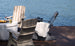 Two Mainstay Adirondack Rockers by Ledge Lounger on a wooden deck face serene water. A nearby fire pit adorned with drinks enhances the scene. One rocker features a striped cushion, and the other is draped in a white blanket, crafting an inviting outdoor oasis for relaxation.