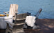 Two Mainstay Adirondack Rockers by Ledge Lounger on a wooden deck face serene water. A nearby fire pit adorned with drinks enhances the scene. One rocker features a striped cushion, and the other is draped in a white blanket, crafting an inviting outdoor oasis for relaxation.