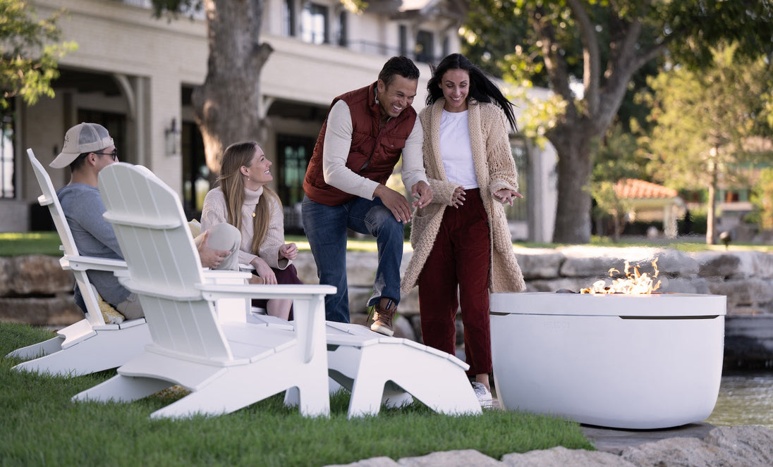 Four people enjoy outdoor relaxation by a fire pit. Two sit on Ledge Loungers white Legacy Adirondack Ottomans while the others stand nearby, engrossed in conversation. Trees and a building backdrop enhance the cozy atmosphere, creating a friendly and relaxed mood.