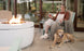 A woman relaxes in a Ledge Lounger Legacy Club Chair on a patio, holding a drink by the fire pit. Dressed in a brown top, white overalls, and slippers, her golden retriever lounges nearby. The outdoor space features customizable design elements and large glass doors in the background.