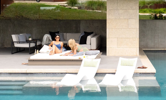Two people relax on a Ledge Lounger Echo Sunbed in a shaded area, surrounded by modern outdoor furniture including a couch and striped cushions. Two white lounge chairs are partially submerged in the pool, enhancing the tranquility of this serene outdoor space.