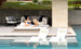 Two people relax on a Ledge Lounger Echo Sunbed in a shaded area, surrounded by modern outdoor furniture including a couch and striped cushions. Two white lounge chairs are partially submerged in the pool, enhancing the tranquility of this serene outdoor space.
