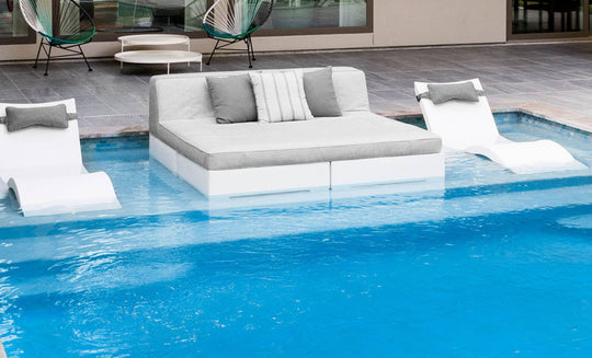 Relax poolside in style with a modern outdoor setup, featuring a Ledge Lounger Affinity Square Sunbed with Backrest Cushion in light gray, centered between two white submerged pool chairs. A chic patio with extra seating and tables completes the backdrop.