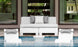 Two Affinity Square Sunbeds with backrest cushions from Ledge Lounger sit partially submerged poolside, facing a modern sofa with gray and white cushions. A small round table nestles between them, while large glass doors reflect the lush greenery beyond.